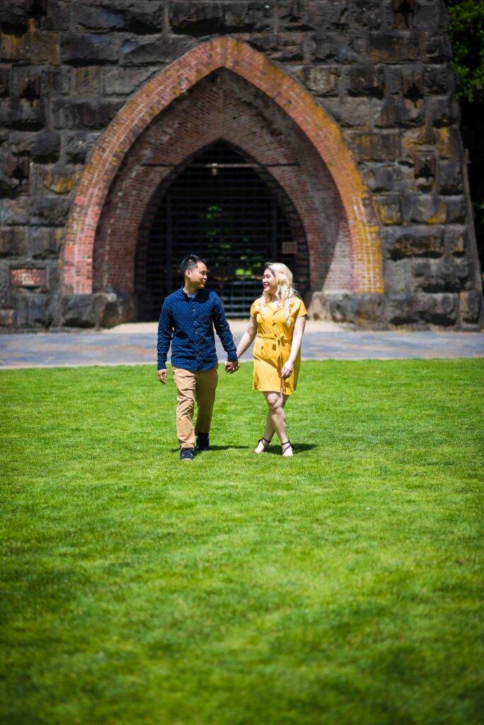 Portrait of an engaged couple walking hand in hand through a grassy area at George Rogers Park in Portland, Oregon. They are smiling at each other in front of the Archway. 