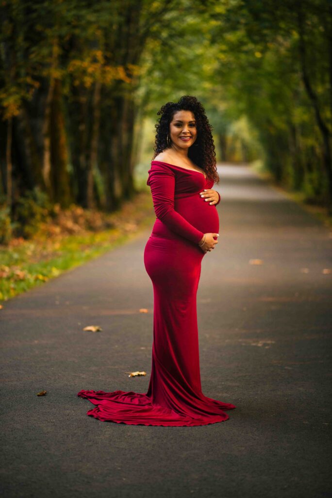 Portrait of a mom-to-be standing on a path at Minto-Brown Island Park in Salem, Oregon. She is holding her baby bump on a tree lined path. 