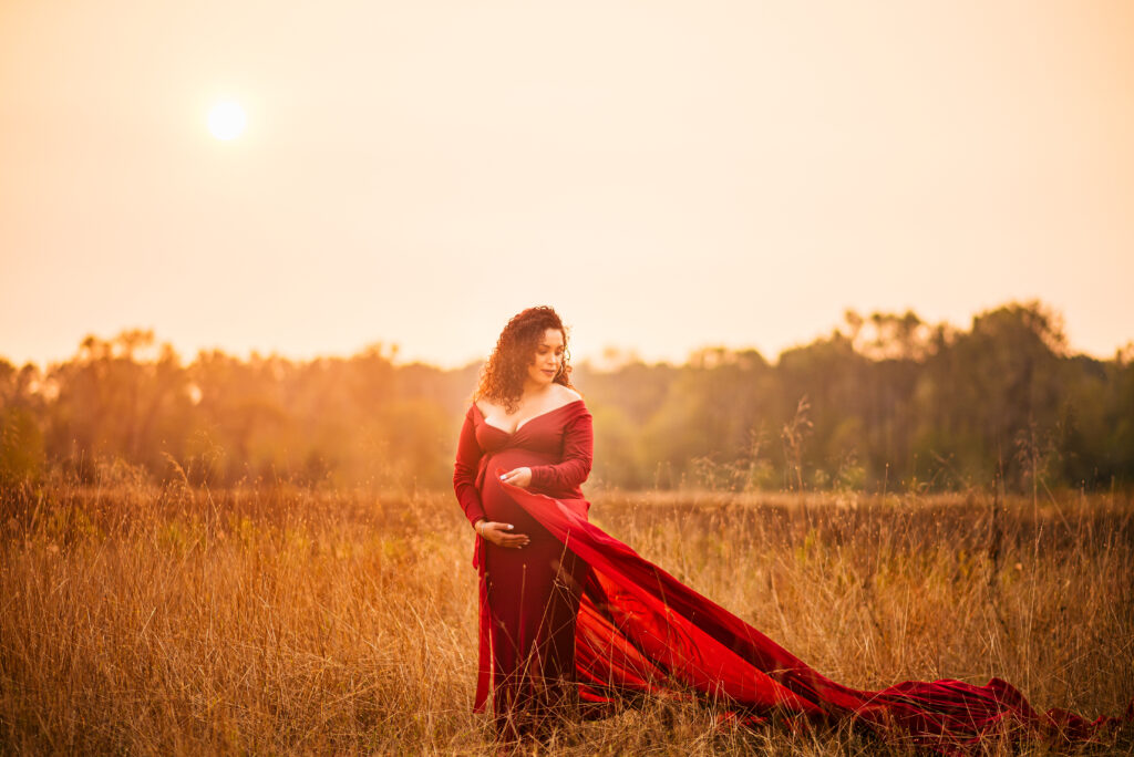 Dreamy golden hour maternity photoshoot of a woman in a red maternity gown at Minto-Brown Island Park in Salem, Oregon. She is holding her dress and looking ethereal in the golden glow of the sun. 