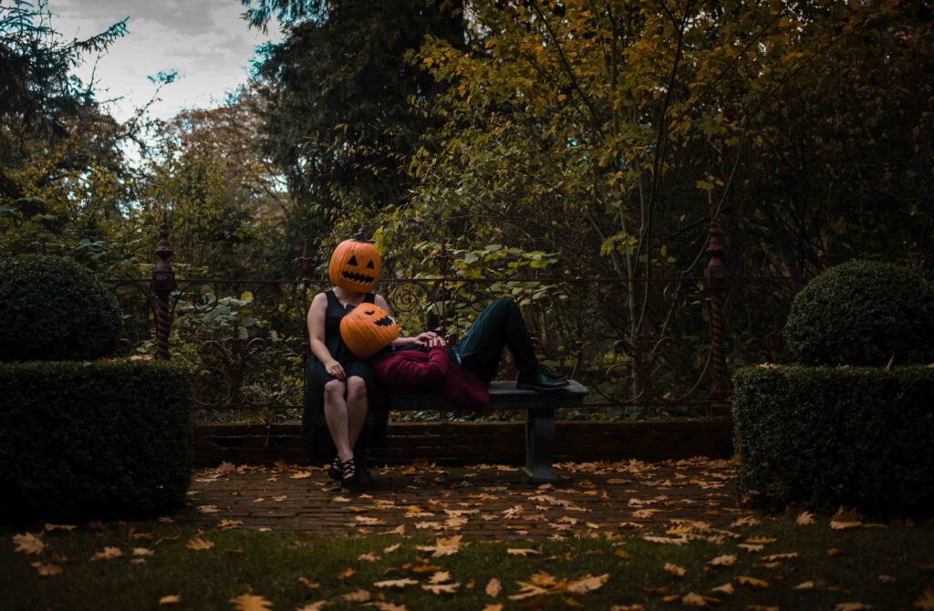 A spooky portrait of a couple wearing pumpkin heads and sitting on a bench in Deepwood Estates and Gardens in Salem, Oregon. The male pumpkin is laying on the bench with his head in the female pumpkins lap next to the sculpted hedges. 