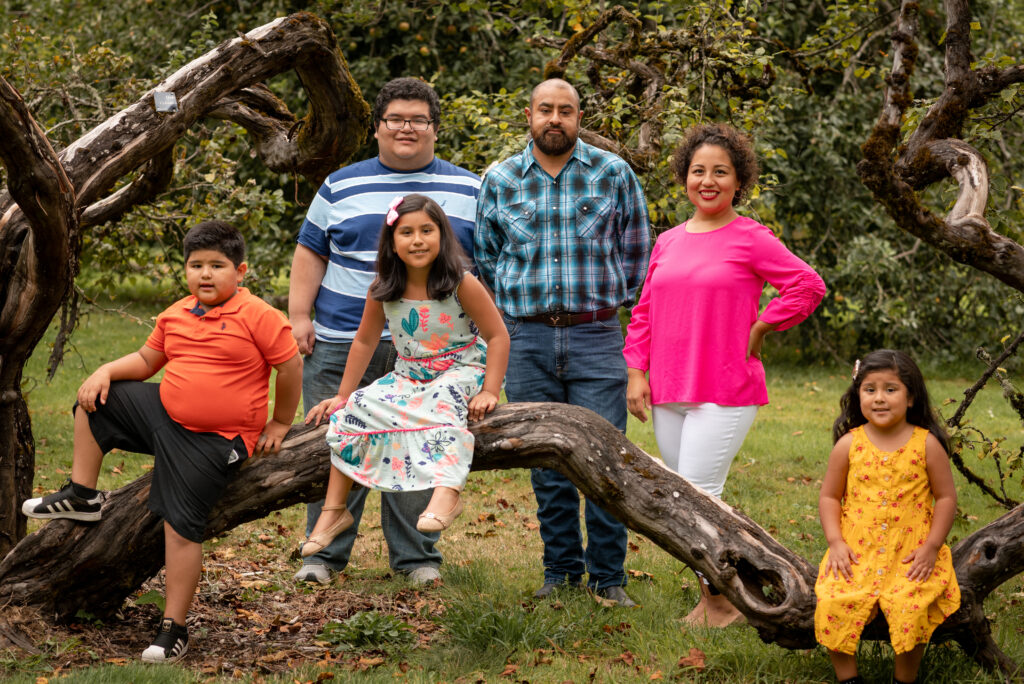 A large family smiles at the camera at Bush's Pasture Park in Salem, Oregon. The children climb on a low horizontal branch while the adults stand behind them. 