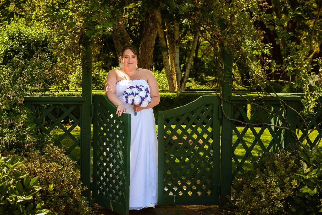 A bride walks through the garden gates at Deepwood Estates and Gardens in Salem, Oregon. She's holding a bouquet of flowers and surrounded by the lush greenery of the garden.