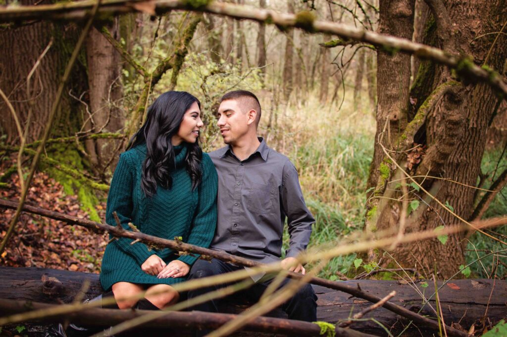 A couple sits together on a log surrounded by forest at Minto-Brown Island Park in Salem, Oregon. They stare lovingly at each other as they share a private moment in the trees. 