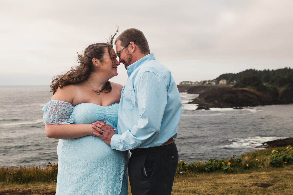 Maternity couple posed on the cliffs at Rocky Creek in Oregon at sunset, with both husband and expecting wife gazing lovingly at each other with a stunning view of the coastline behind them