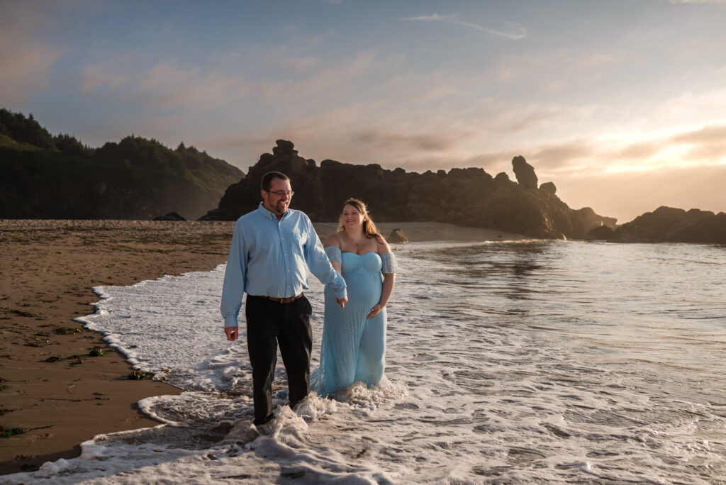 Maternity couple walk along the waters edge at Fogarty Beach in Oregon at sunset, with both husband and wife holding hands and laughing as the sunsets behind them