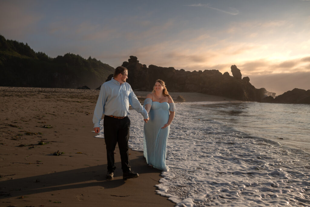 Maternity couple walk along the waters edge at Fogarty Beach in Oregon at sunset, with both husband and wife holding hands and looking at each as the sunsets behind them