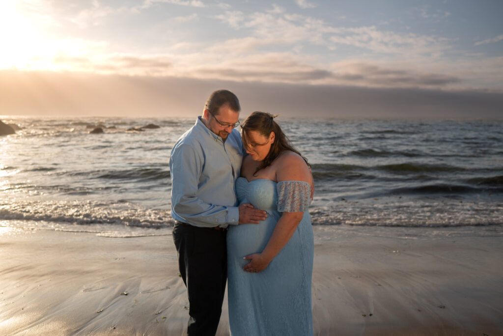 Maternity couple walk along the waters edge at Fogarty Beach in Oregon at sunset, with both husband and wife staring lovingly at the baby bump with the sunsetting on the water behind them