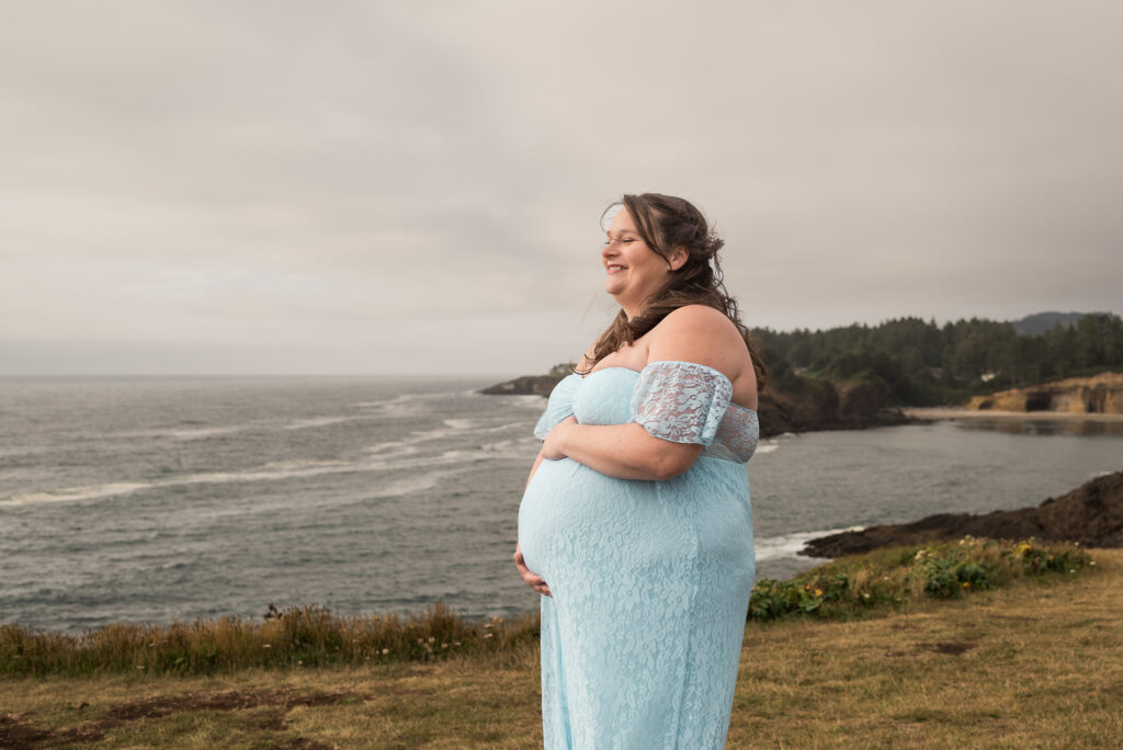 Expectant mom holds her baby bump on the cliffs at Rocky Creek in Oregon at sunset with a stunning view of the coastline behind her. 