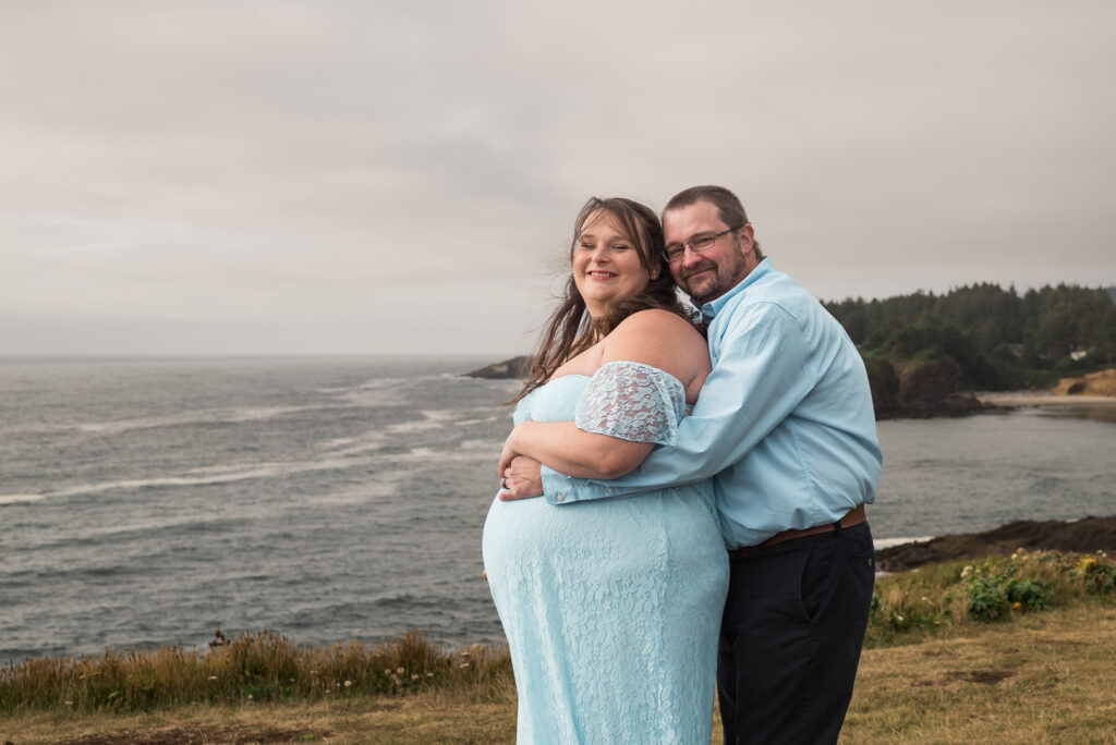 Maternity couple posed on the cliffs at Rocky Creek in Oregon at sunset, with both husband and expecting wife embracing with a stunning view of the coastline behind them